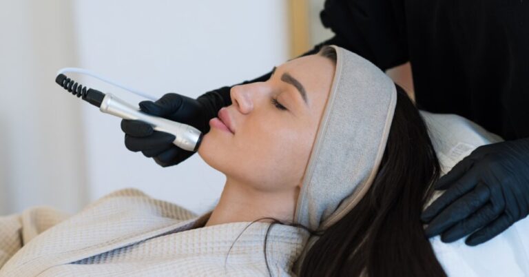 A woman lying down with her eyes closed while a person wearing black gloves uses a handheld wand device on her jawline during a facial treatment