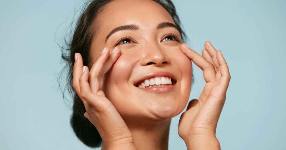 A smiling woman with a bright, glowing complexion looking slightly upward while lightly touching her cheeks with both hands against a light blue background
