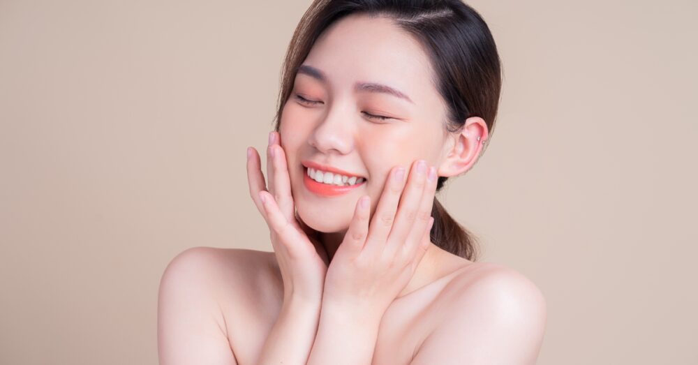 A woman with clear, glowing skin touching her face and smiling against a neutral background
